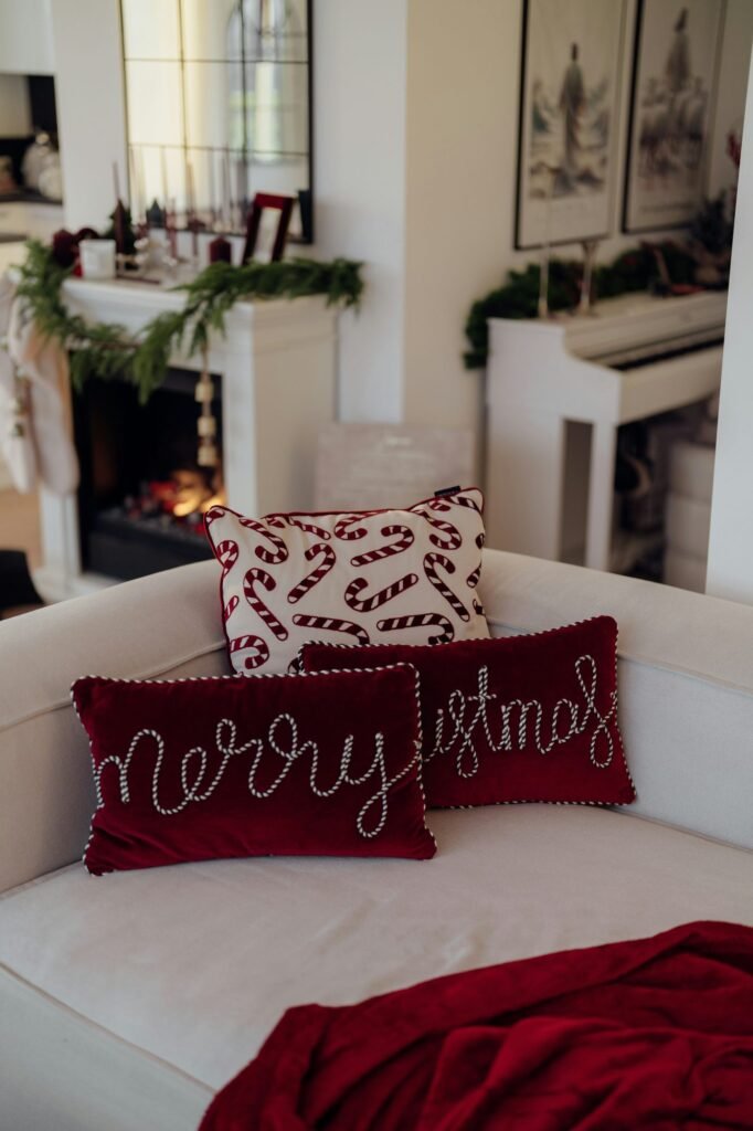 Sofá blanco con cojines rojos bordados con mensajes navideños junto a una chimenea decorada