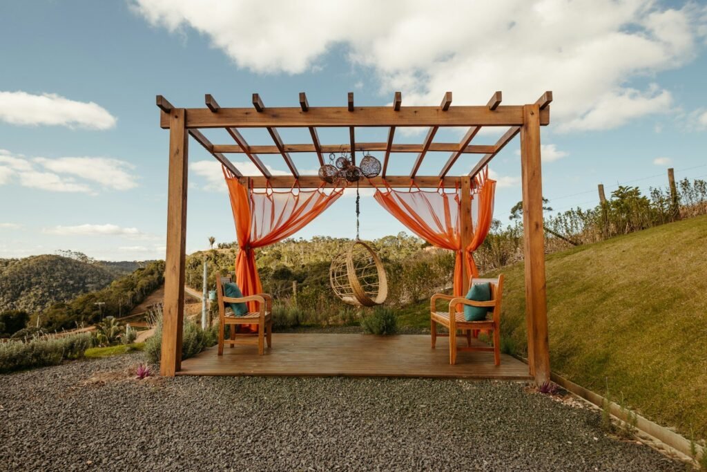 Pérgola de madera con cortinas naranjas y sillas frente a un paisaje de colinas